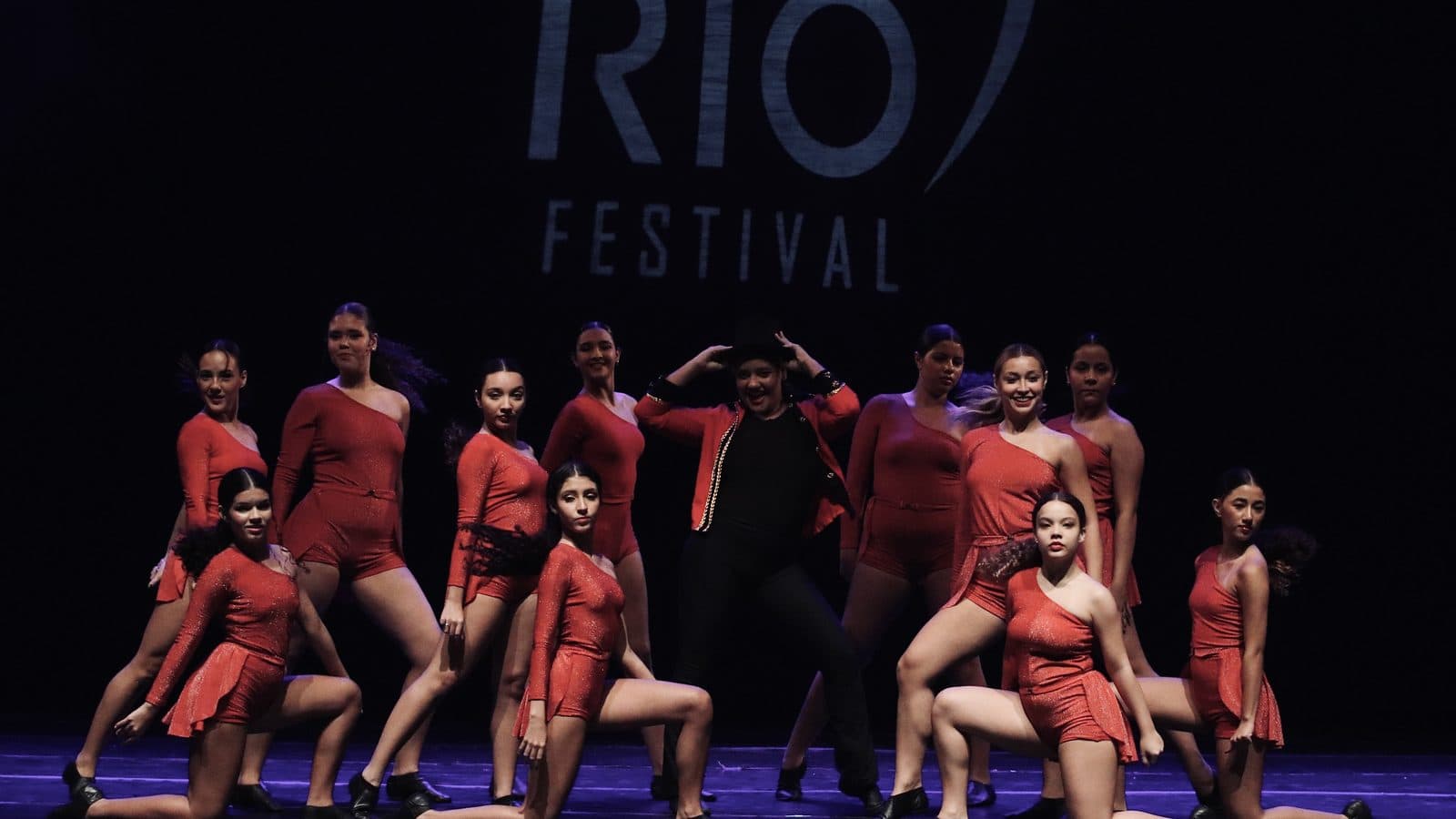 Grupo de bailarinas de figurino vermelho em pose final no palco, com o logo do MoviRio Festival ao fundo — Mostra Competitiva 2026
