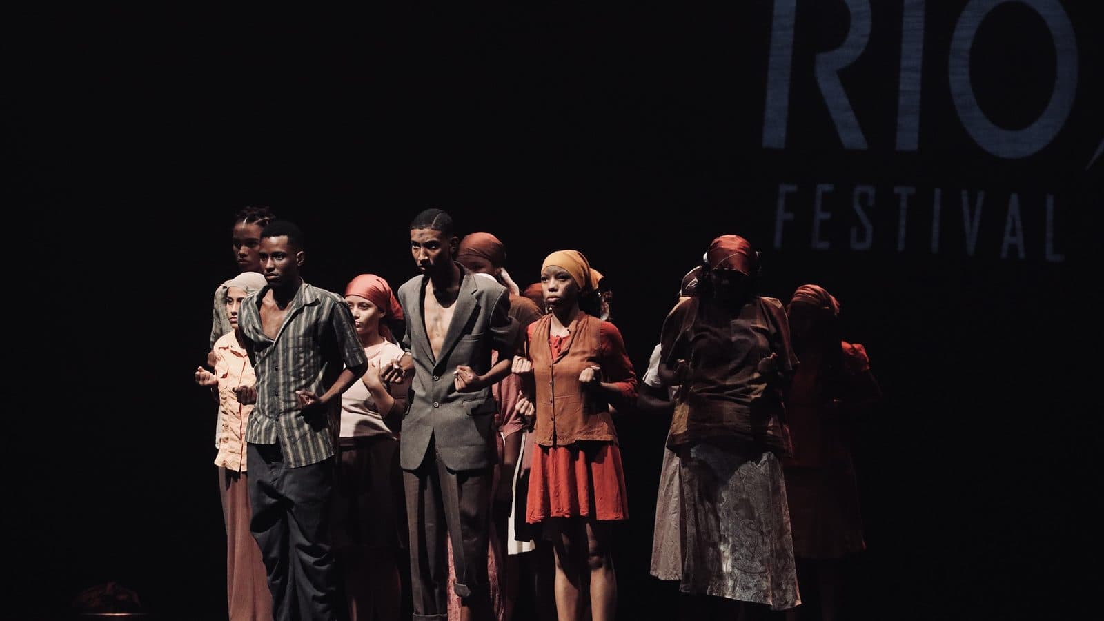 Elenco em figurinos de época reunido no palco em cena de dança, com o logo do MoviRio Festival ao fundo — Mostra Competitiva 2026
