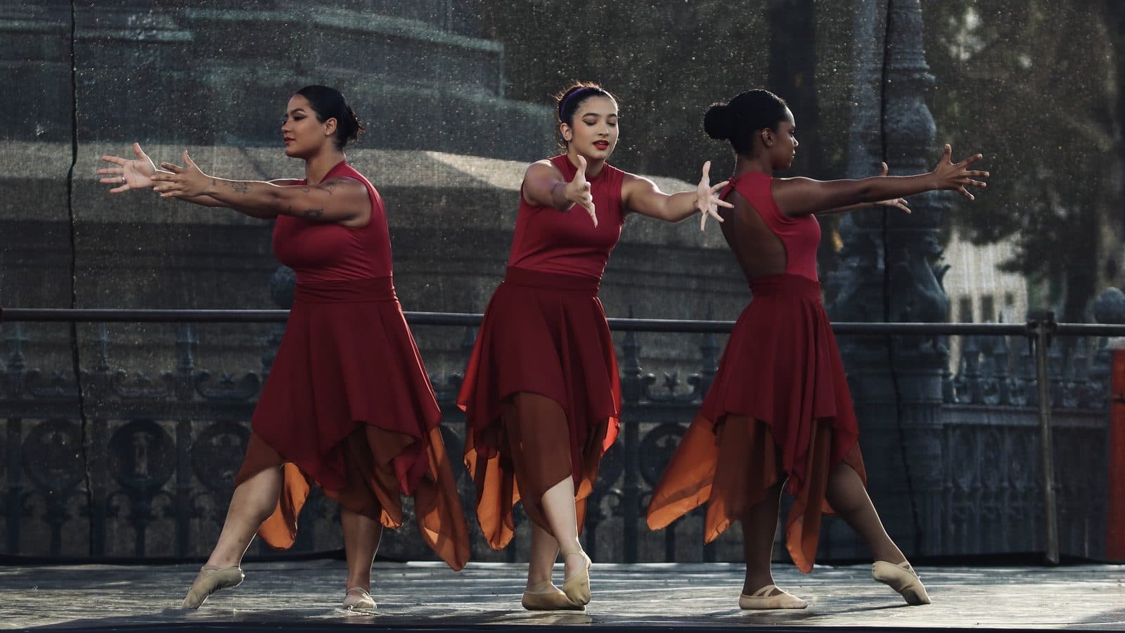 Três bailarinas de vestido vermelho dançando no Palco Rio, ao ar livre, com o monumento da Praça Tiradentes ao fundo — MoviRio 2026